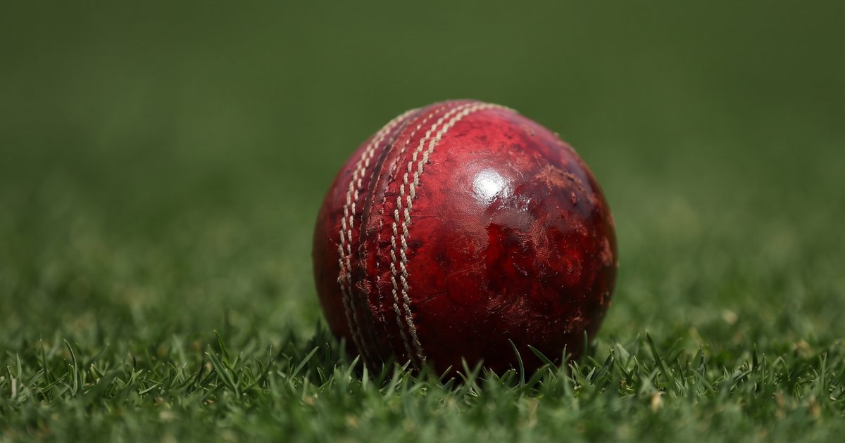 Close-up of a worn red cricket ball on the outfield, one side scuffed and one side polished